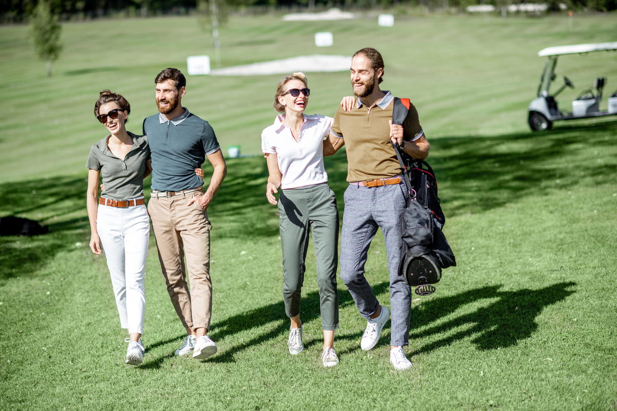 Vier glückliche Menschen, zwei Männer und zwei Frauen, spazieren gemeinsam über einen grünen Golfplatz. Sie lachen und wirken entspannt, während einer der Männer eine Golftasche trägt. Im Hintergrund ist ein Golfwagen zu sehen, der die ruhige und freundliche Atmosphäre des Golfplatzes ergänzt.