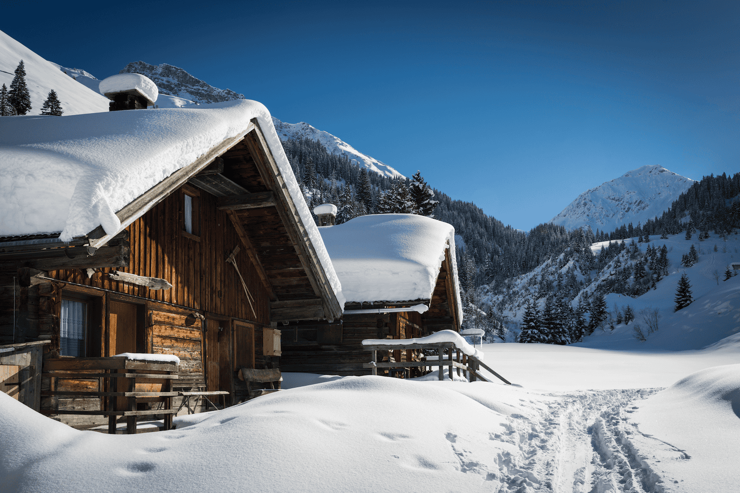 Verschneite Berghütten aus Holz in einer idyllischen Winterlandschaft, umgeben von schneebedeckten Bergen und dichten Tannenwäldern. Der klare blaue Himmel strahlt über der Szene, während Spuren im Schnee zur Hütte führen und ein Gefühl von Ruhe und Abgeschiedenheit vermitteln. Perfekt für einen erholsamen Winterurlaub inmitten der Natur.